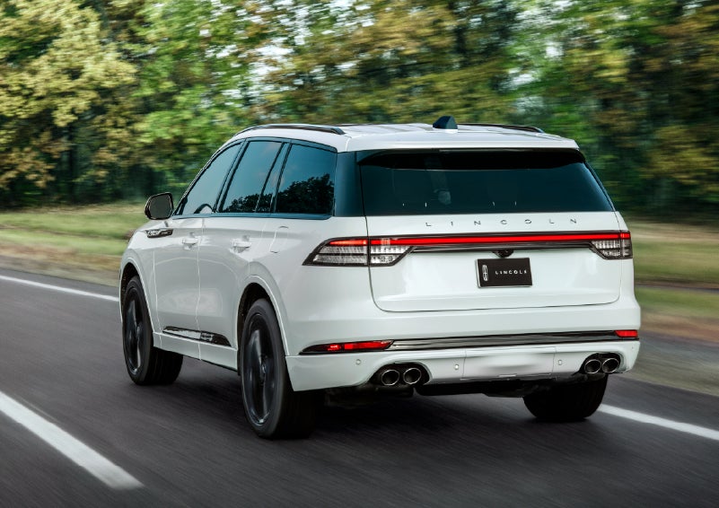 A white 2026 Lincoln Aviator® SUV is being driven away down a road lined with colorful trees. | Lincoln of Troy in Troy MI