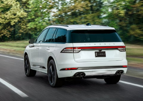 A white 2026 Lincoln Aviator® SUV is being driven away down a road lined with colorful trees. | Lincoln of Troy in Troy MI