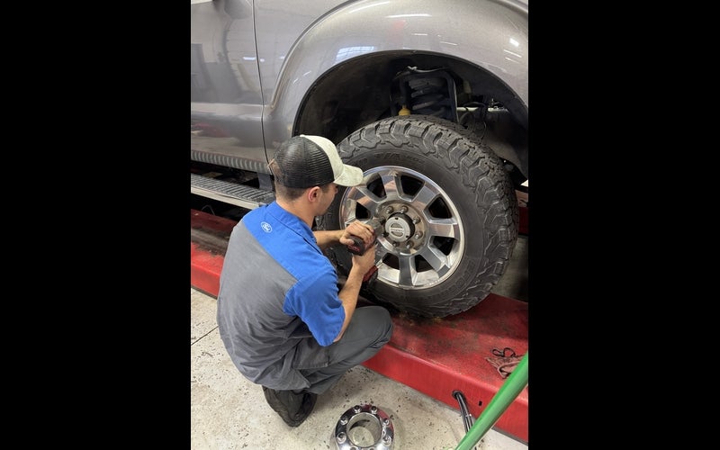 A person kneels beside a raised gray pickup truck in an automotive workshop