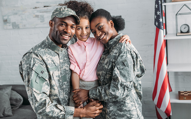 Three people standing together and embracing in a home setting; two are wearing camouflage uniforms