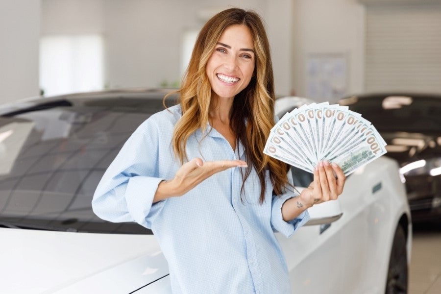 Person standing in a car showroom, holding a fan of U.S. dollar bills and gesturing toward them, with a white vehicle in the background