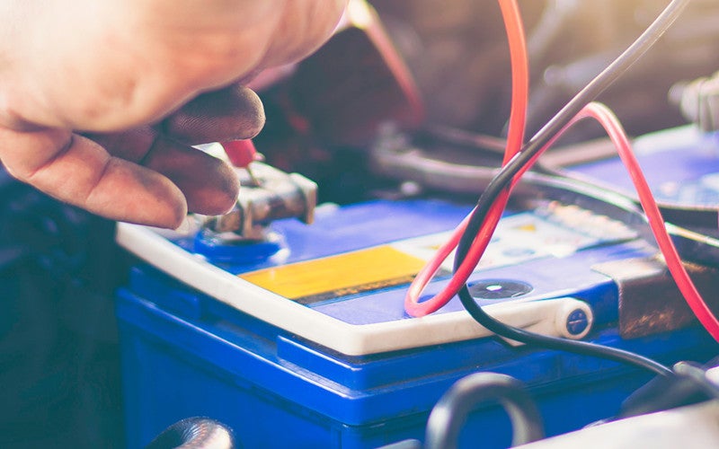 Hand connecting red and black wires to the terminals of a blue car battery