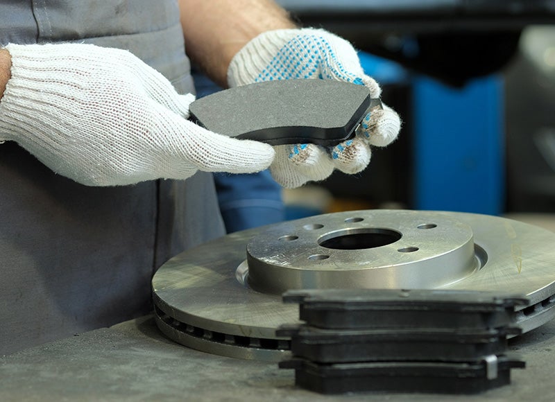 Close-up of gloved hands holding a brake pad above a metal brake rotor, with additional brake pads stacked
