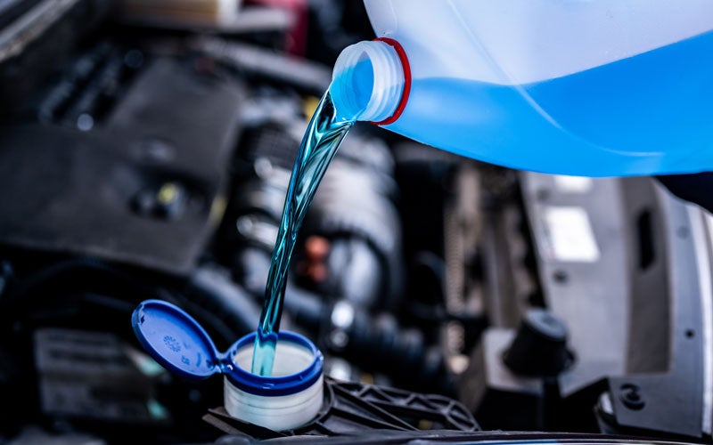Blue windshield washer fluid being poured from a plastic container into a vehicle washer fluid reservoir under the hood of a car