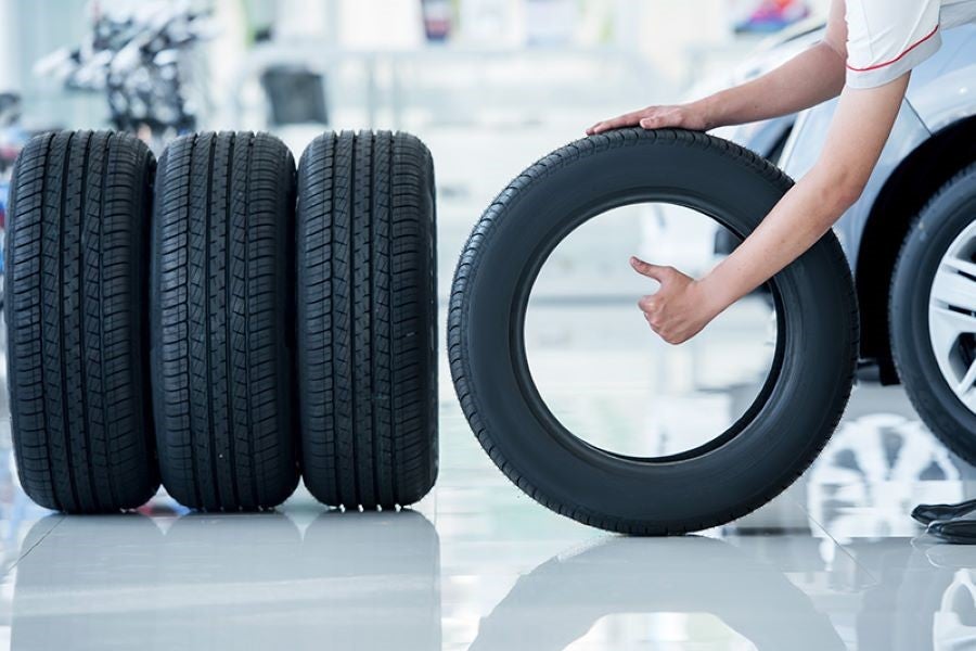 Person holding a car tire with a thumbs-up, next to three other new tires and a car in a bright shop